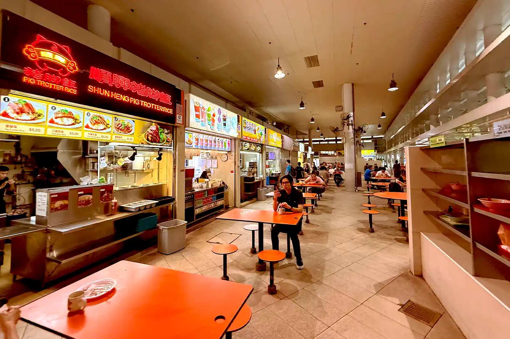 A wide-angle view of Bedok Interchange Hawker Centre during a quiet hour, featuring rows of food stalls with glowing signs and patrons dining at orange tables.