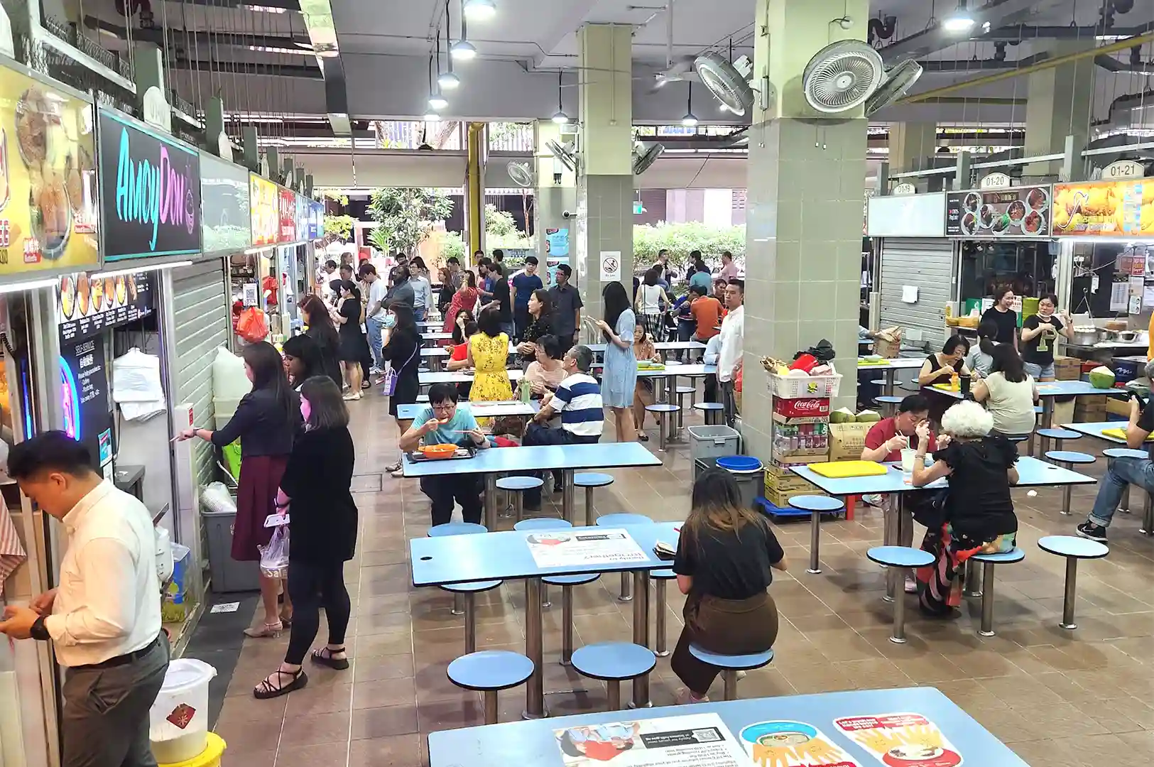 A bustling hawker centre in Singapore with people dining at tables and queuing at food stalls, capturing the lively atmosphere before closing time.
