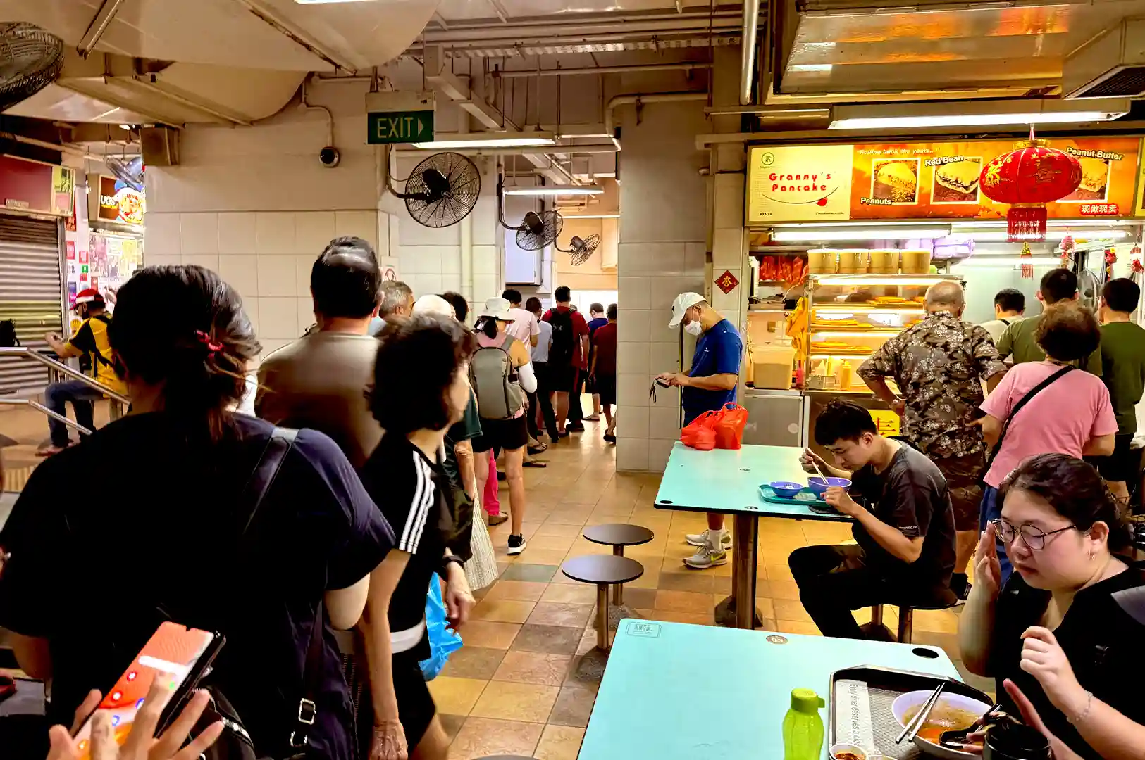 A candid street-style photograph capturing the quiet, everyday moments of a bustling Singapore hawker centre, featuring patrons dining at turquoise tables, lines forming at food stalls like Granny's Pancake, and slow-paced movement that defines local hawker culture.