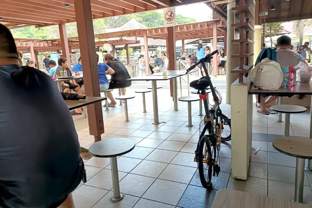 A quiet corner of East Coast Lagoon Food Village showing diners seated under a wooden pavilion next to a parked bicycle, capturing a peaceful moment before the table changes.
