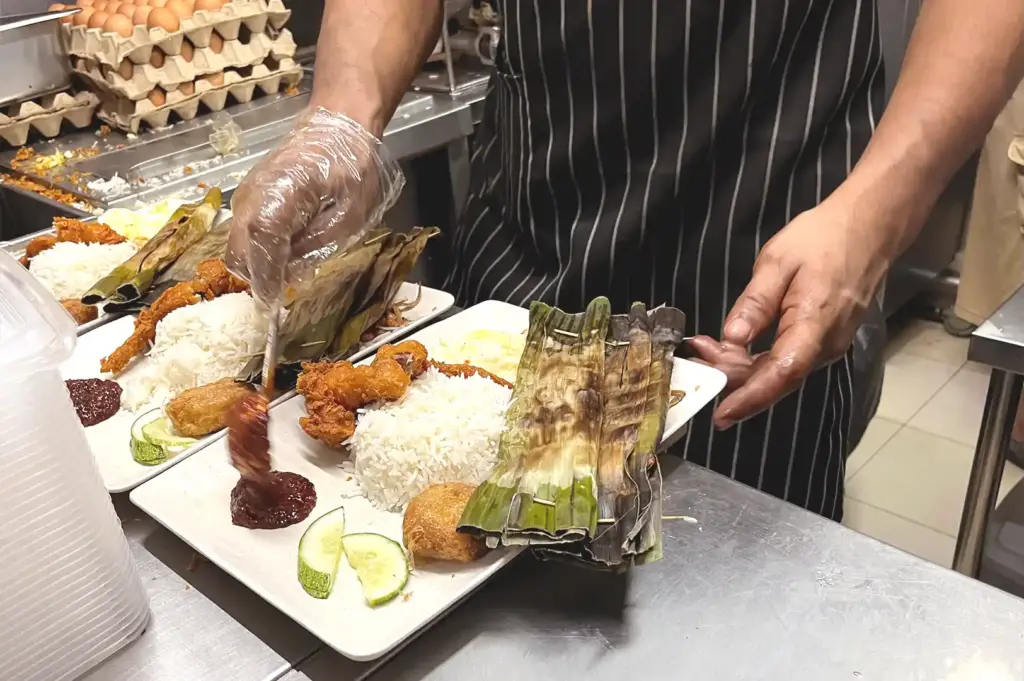 A hawker center chef wearing a striped apron and plastic gloves carefully plating traditional rice dishes wrapped in banana leaves with sambal chilli at Adam Road Food Centre.