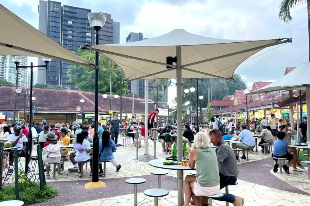 A crowded outdoor dining plaza featuring large shade umbrellas, tables filled with patrons, and urban high-rises in the background during peak dining time.