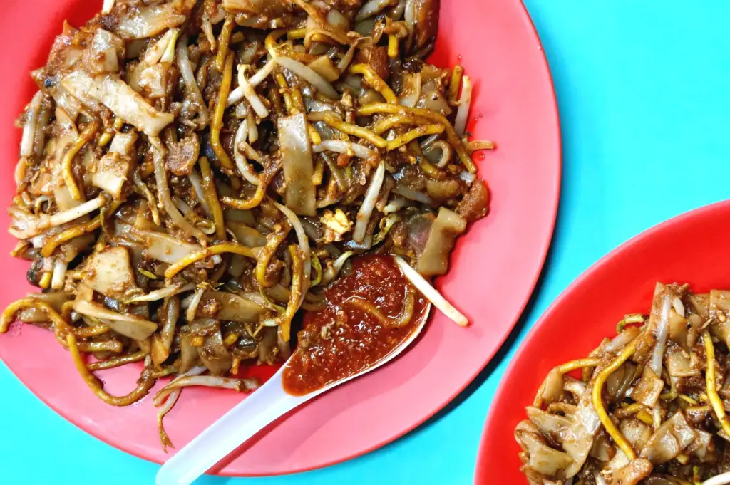 Top-down view of char kway teow served on a red plate with a white spoon holding spicy chilli paste at Hong Lim Food Centre hawker lunch.