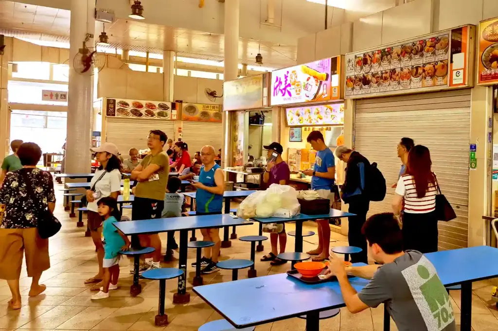 Patrons and families gathering around blue tables at Bedok Interchange Hawker Centre, waiting in line and enjoying meals during an ordinary afternoon.
