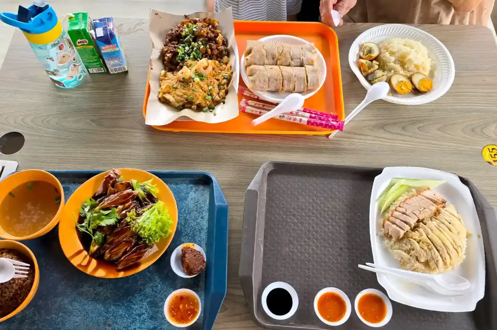 A vibrant spread of local hawker dishes—including fried carrot cake, popiah, chicken rice, and roasted duck—laid out across trays on a wooden table, reflecting shared meals and repeated movements.