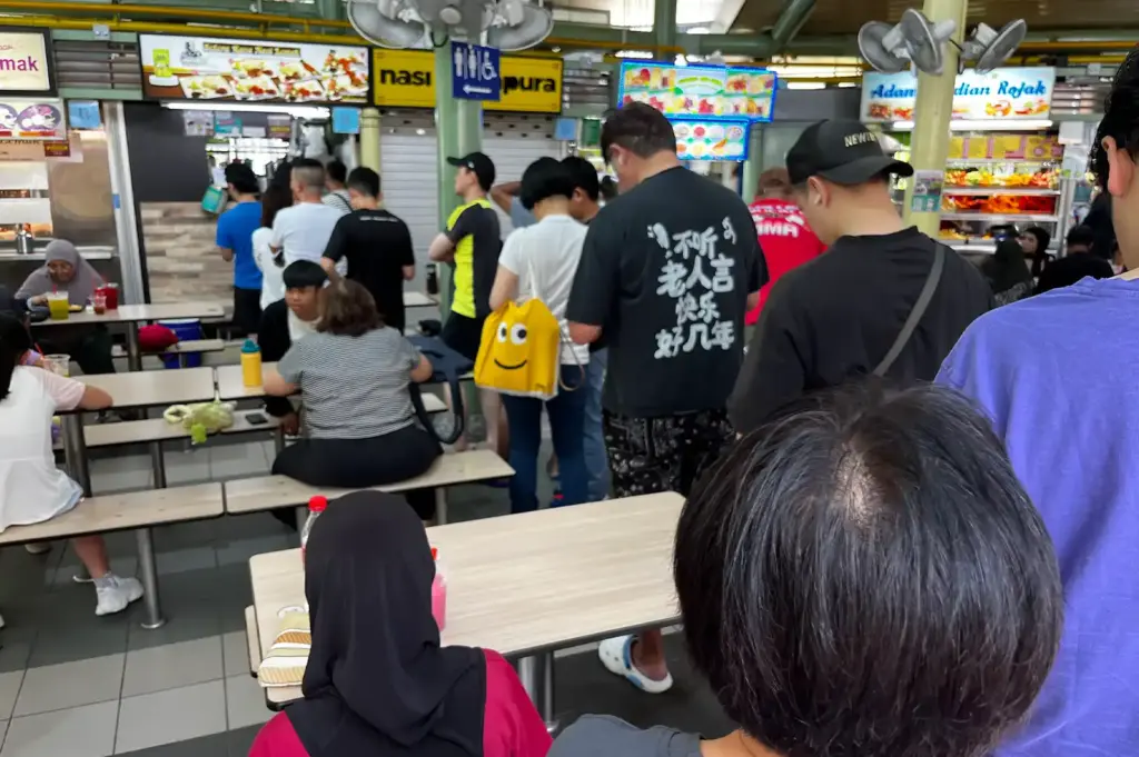 A close-up view of a queue and seating area inside Adam Road Food Centre, capturing the lively dining atmosphere and familiar routines of local food culture.