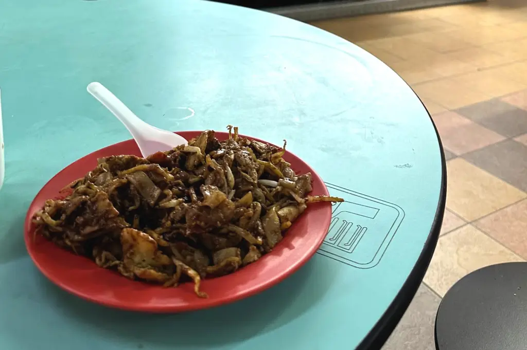 A plate of traditional char kway teow with a white plastic spoon resting on a turquoise hawker table at Hong Lim Food Centre during lunchtime.