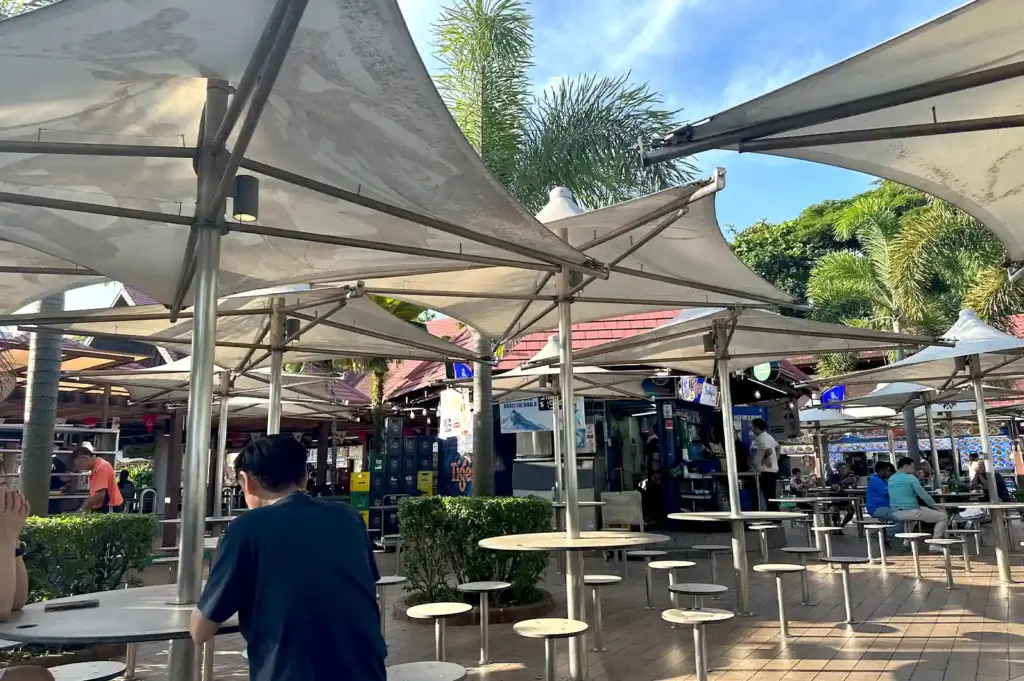 Large white shade sails arching over the open-air dining area at East Coast Lagoon Food Village under a blue sky, framing a quiet afternoon observation of hawker culture.