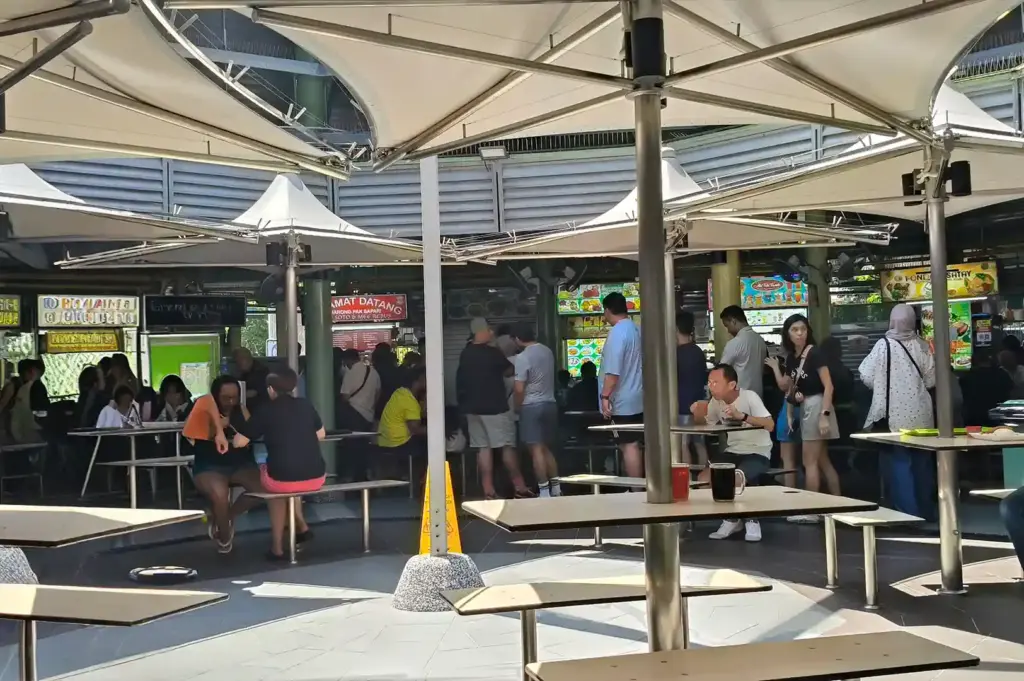 atrons dining under large white shade structures at Adam Road Food Centre, showcasing the morning and afternoon rhythms of a popular Singapore hawker space.