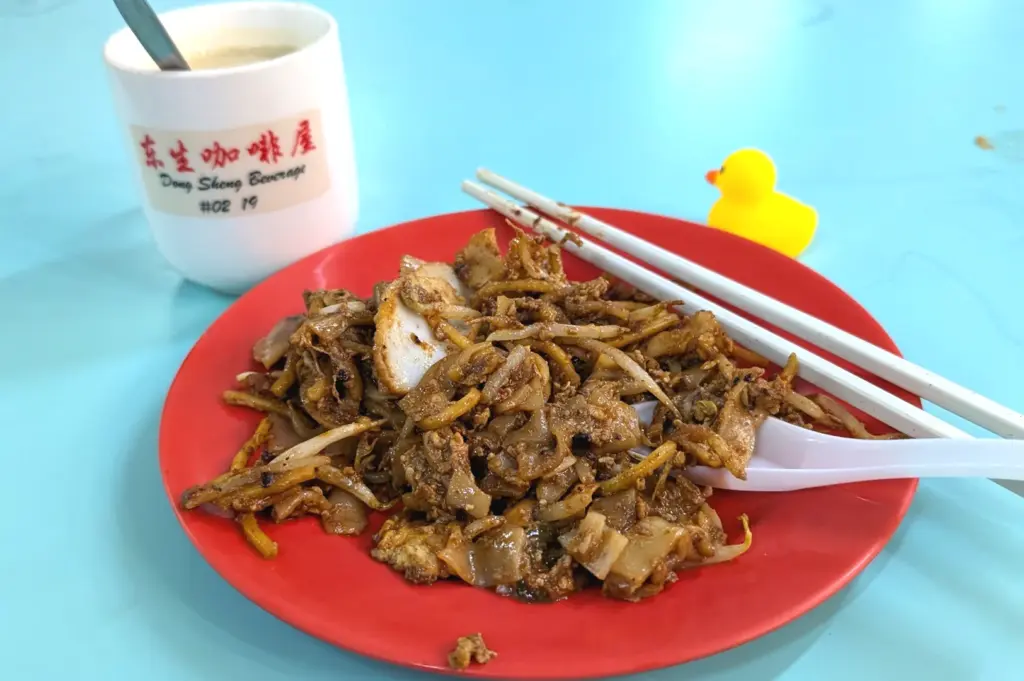 Char kway teow served on a red plate alongside a cup of local coffee and chopsticks at Hong Lim Food Centre, capturing a vibrant hawker meal.
