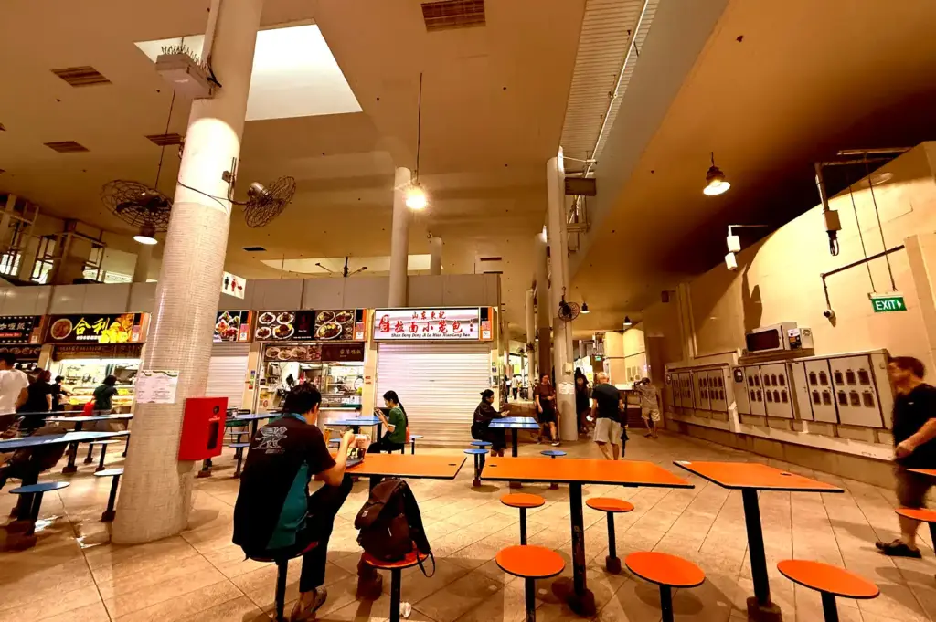 A quiet aisle inside Bedok Interchange Hawker Centre showing concrete pillars, closed shutter stalls, and patrons seated at orange tables.