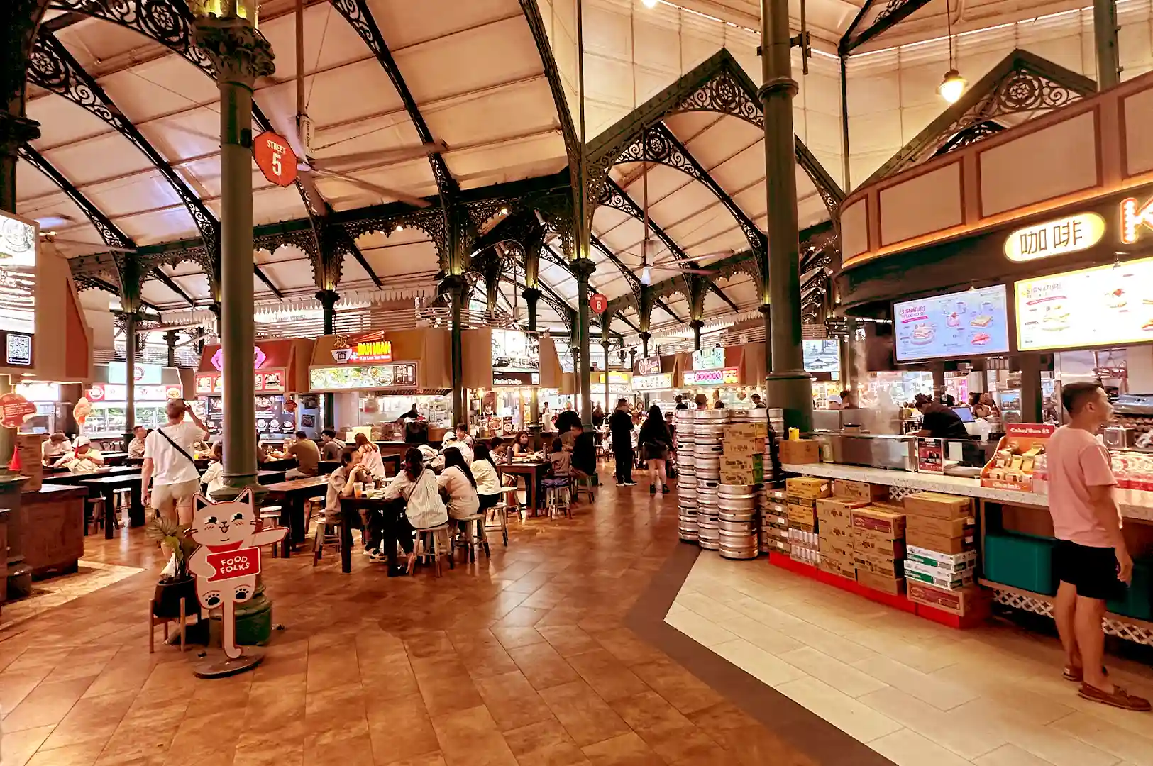A bustling, high-ceilinged Singapore hawker center featuring rows of food stalls, iron pillars, and diners seated at communal tables, capturing the familiar journeys and lively atmosphere tied to favorite local meals.