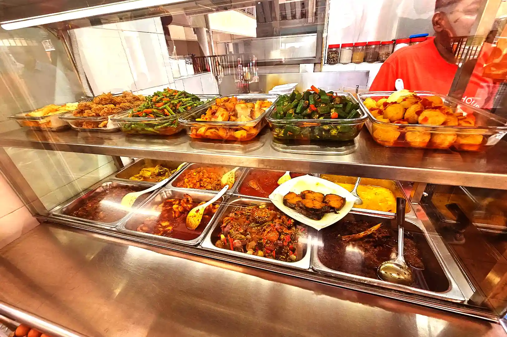 A vendor preparing dishes behind a display glass at a hawker centre food stall.