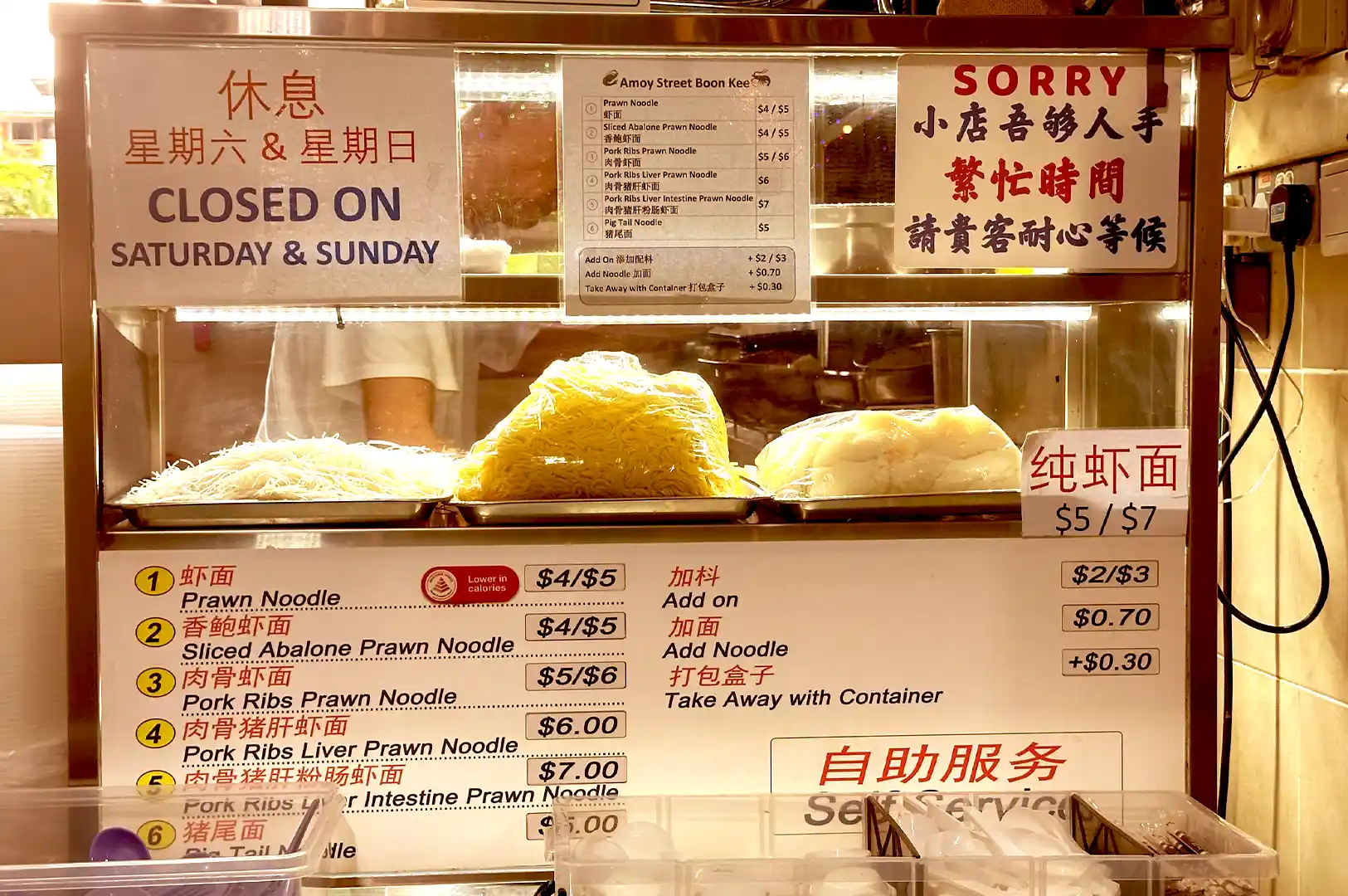 A display case at a hawker stall showing fresh yellow noodles and bee hoon, with bilingual menu boards and signs detailing prices and operating hours.