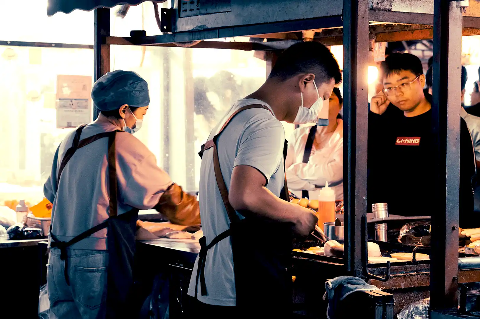 A nighttime view of hawker stall chefs wearing masks and aprons working inside a bustling open-air kitchen stall, illuminated by warm overhead lights and a gas cylinder in the foreground.