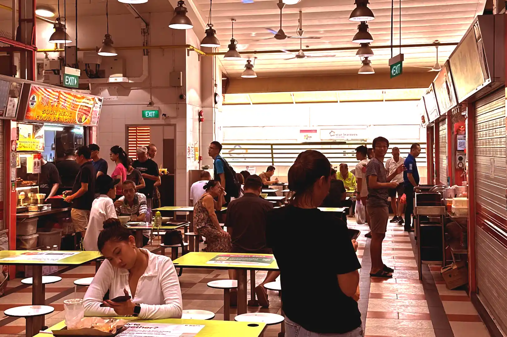A bustling indoor hawker centre in Singapore featuring patrons dining at yellow tables, food stalls with bright signs, and people engaged in everyday routines.