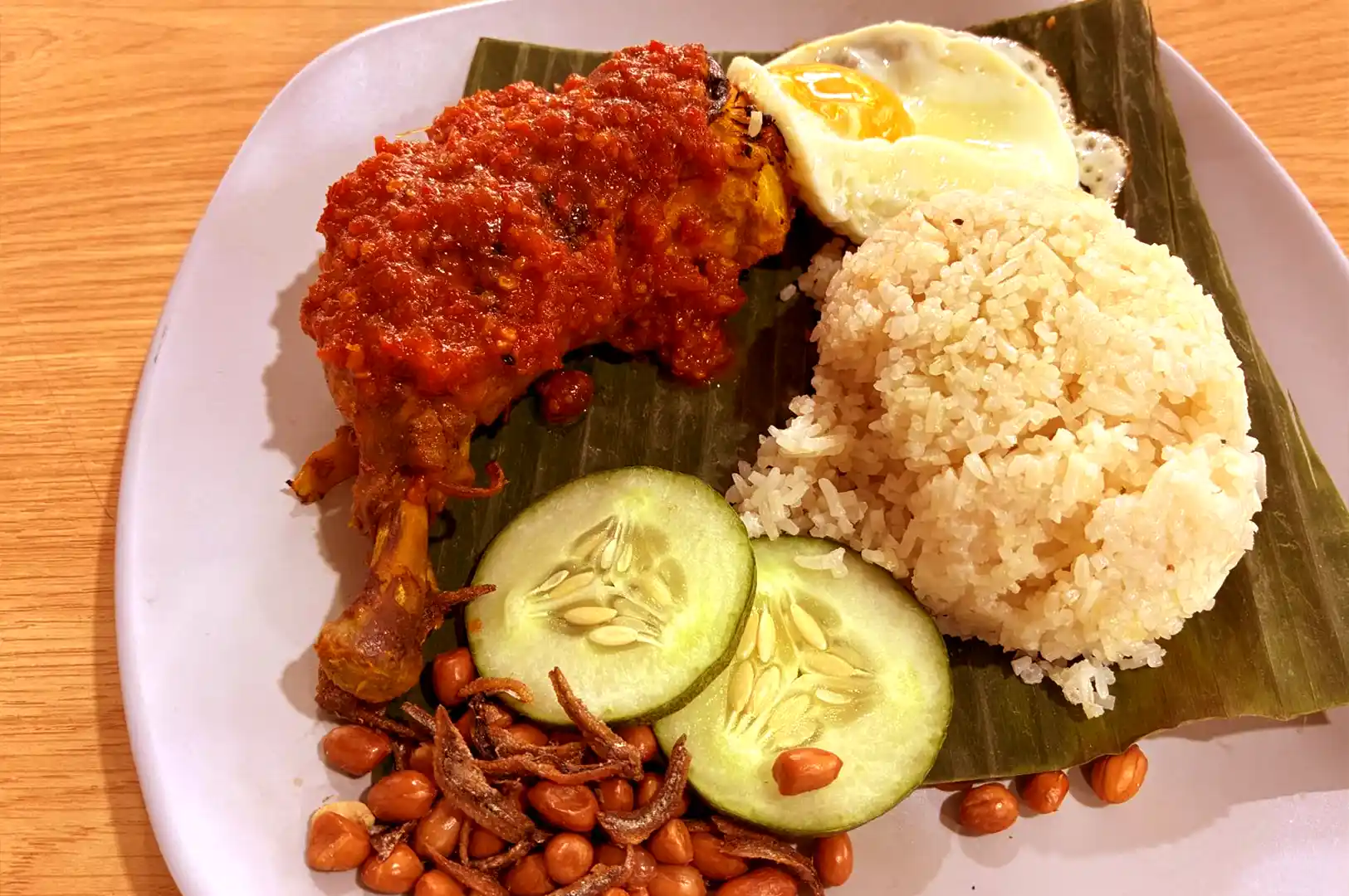 A plate of nasi lemak served on a banana leaf featuring rice, a fried chicken leg generously coated in bright red sambal, a sunny-side-up egg, cucumber slices, peanuts, and anchovies.