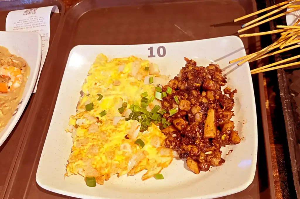 A white square plate featuring a half-and-half portion of savory fried carrot cake (chai tow kway)—one side light and eggy, the other dark and sweet—served alongside satay skewers, capturing the comforting memories of returning to beloved hawker stalls.