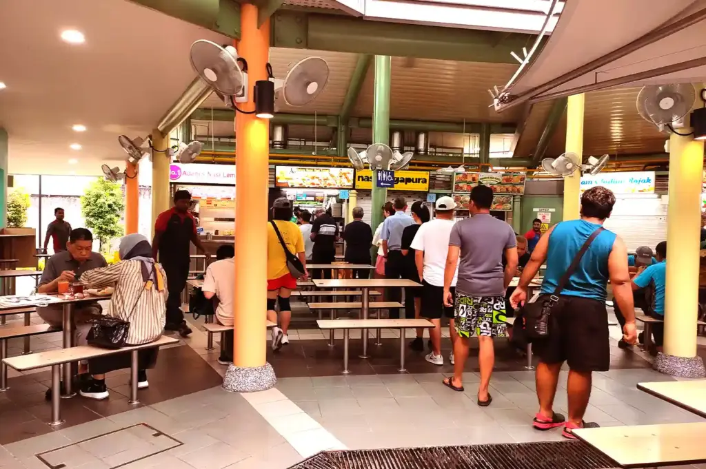 A bustling open-air hawker centre dining area filled with people queuing at food stalls and seated at tables.