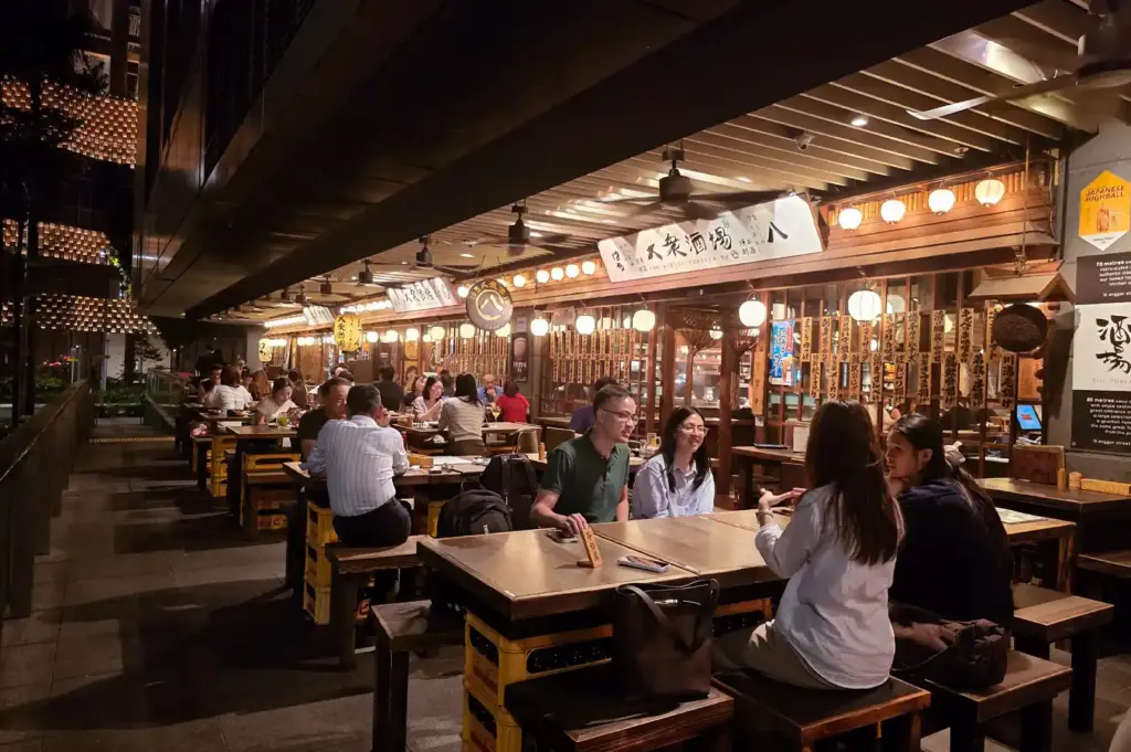 A bustling outdoor dining area at an izakaya featuring lantern-lit spaces, patrons enjoying shared dishes, and vibrant Japanese pub culture in Singapore.