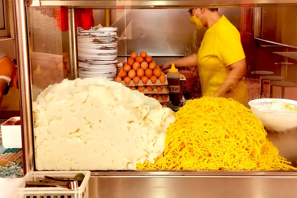 Inside a hawker stall glass counter, mounds of yellow noodles and white rice noodles sit next to stacks of plates, a carton of eggs, and a small crate of limes.