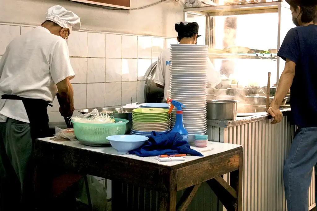 A wooden hawker stall preparation counter featuring scratches, stains, and worn surfaces alongside towering stacks of ceramic plates, colorful bowls, and a Thai menu sign on the wall, quietly revealing years of repeated work.