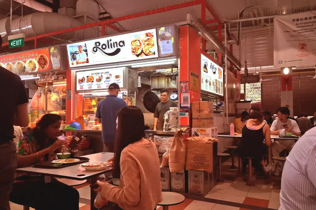 A vibrant food stall at a local hawker centre, showing customers enjoying meals at tables and staff working inside the kitchen.