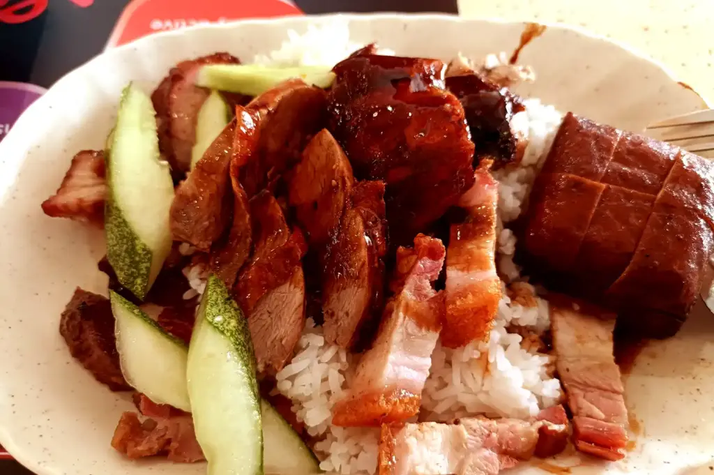A plate of hainanese roasted chicken rice served over rice with sliced cucumbers, highlighting the savory dishes that arrive on the table during a shared meal.