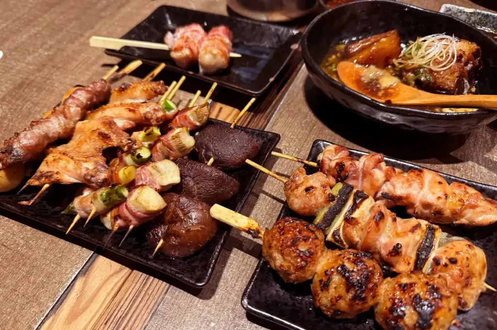 A close-up of shared izakaya dishes featuring grilled yakitori skewers, bacon rolls, shiitake mushrooms, and a savory bowl of braised meat.