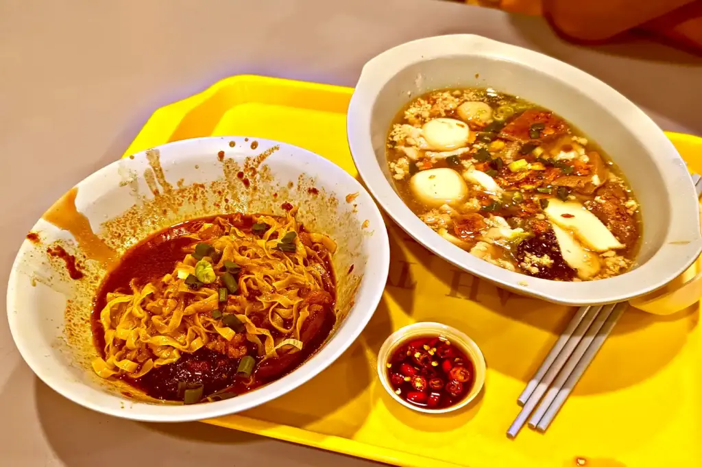 A yellow tray on a table featuring a bowl of dry noodles in rich sauce, a bowl of prawn noodle soup with ingredients like fishballs and sliced meat, and a small dish of sliced chili.