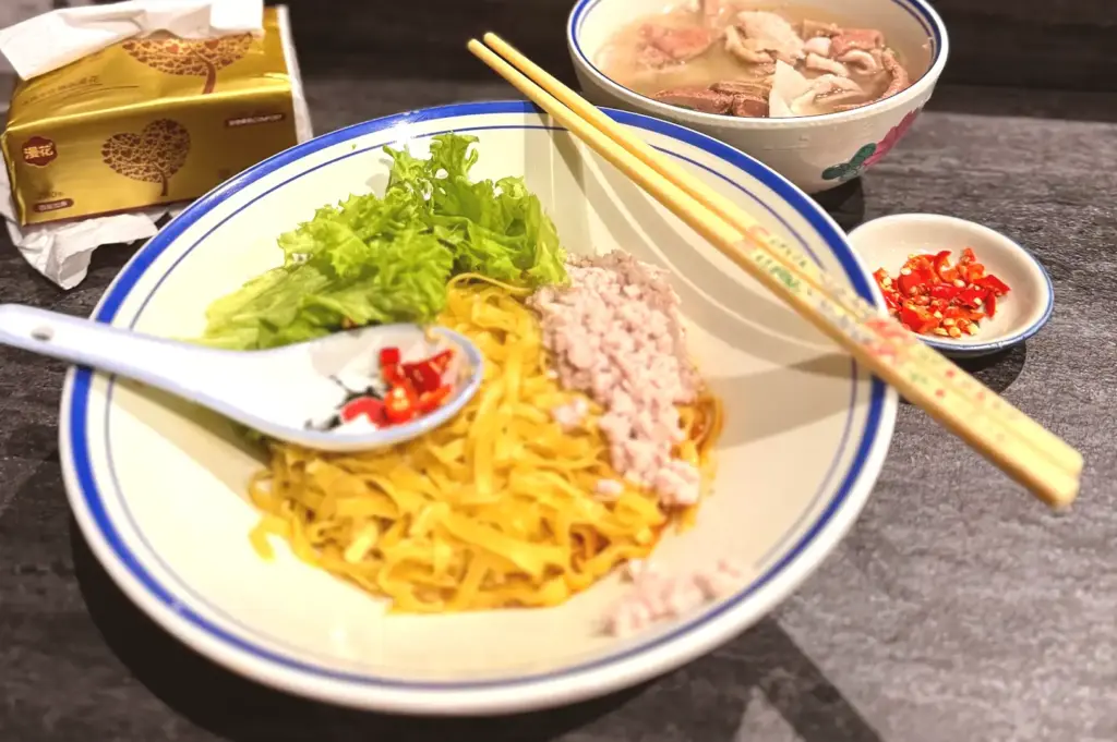 A bowl of dry-style bak chor mee noodles accompanied by a pair of wooden chopsticks, a porcelain spoon, and a small dish of sliced red chilies.