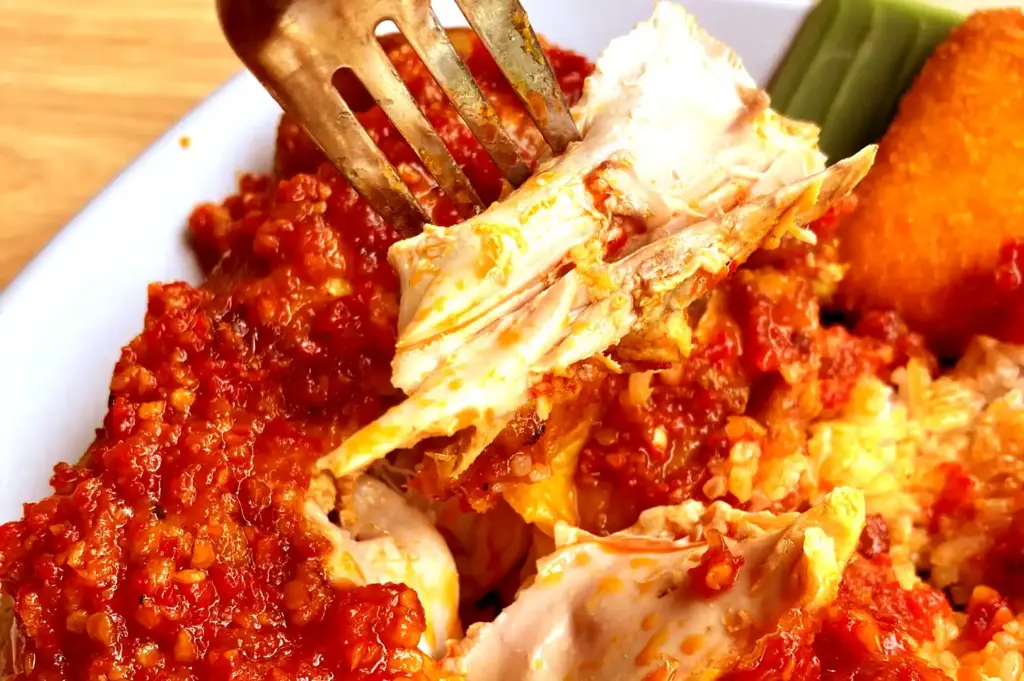 A fork lifting a tender piece of chicken covered in spicy sambal from a nasi lemak dish, capturing the intimate, changing moments of a shared meal.