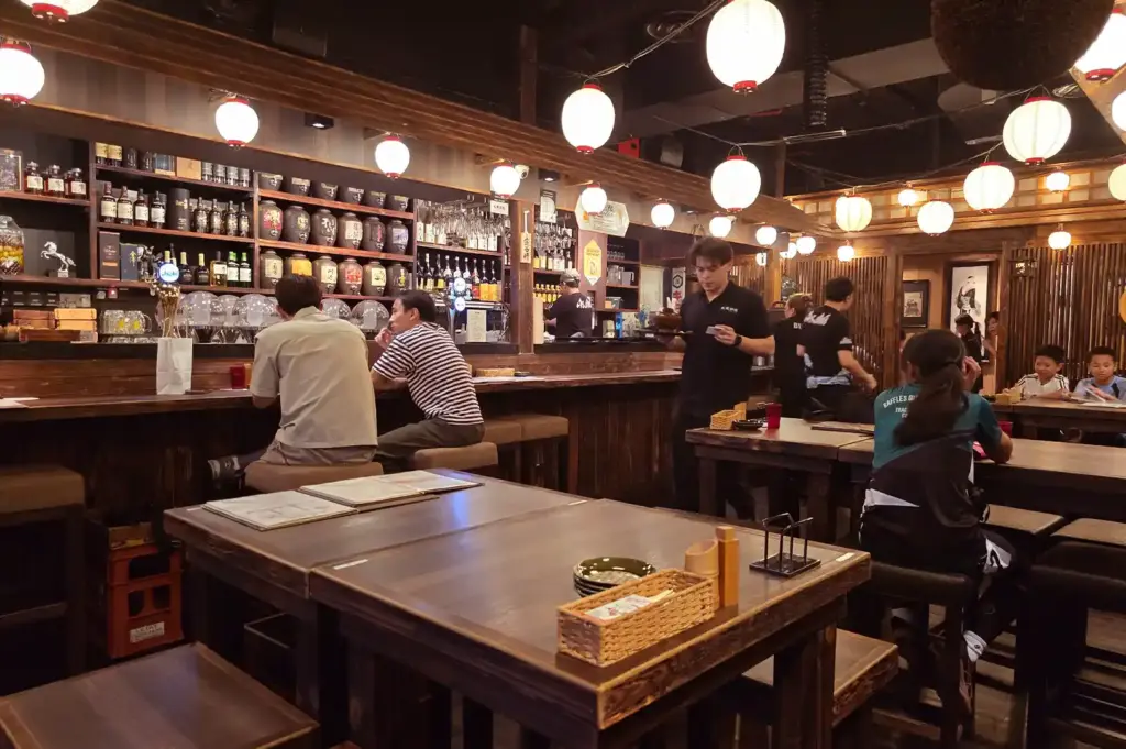 The cozy interior of a traditional izakaya with warm paper lanterns, a fully-stocked bar counter, and quiet moments captured during evening dining.
