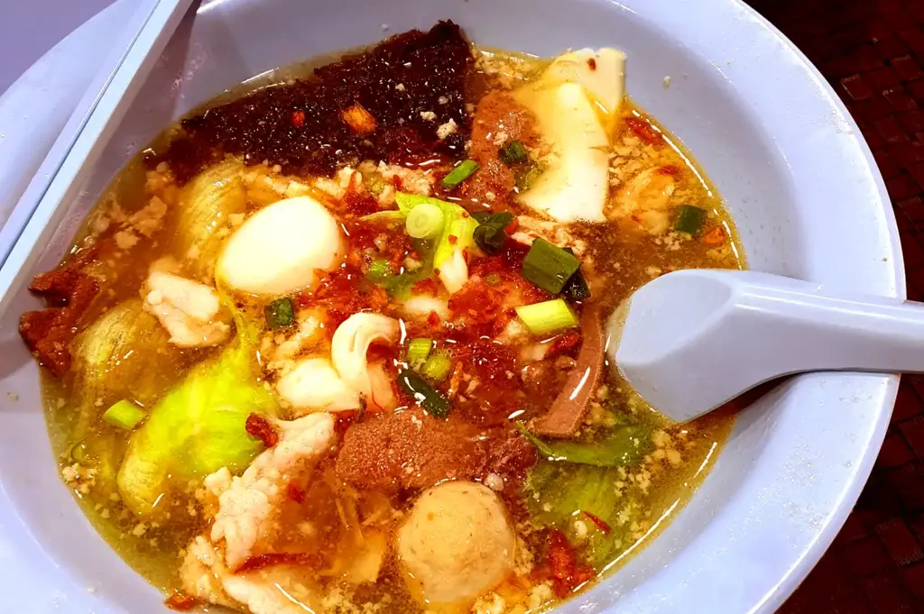 A close-up view of a blue bowl filled with a savory prawn soup containing noodles, meatballs, pork slices, and greens, accompanied by a matching spoon and chopsticks.