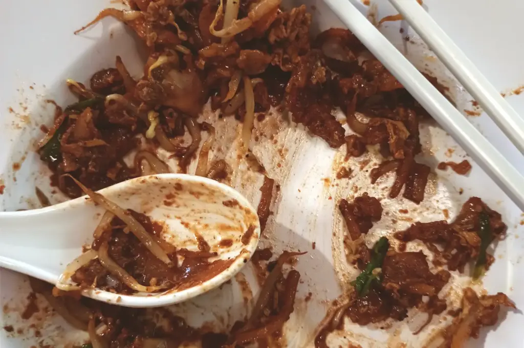A half-eaten plate of char kway teow showing remnants of the meal, complete with a white spoon and chopsticks resting on the side.