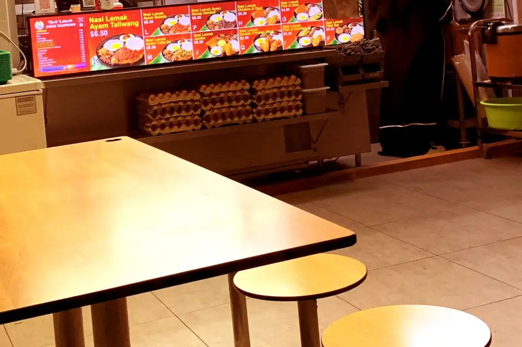 A quiet dining table with wooden stools set in front of a food stall counter displaying a bright menu and stacks of egg trays, setting the scene where local dishes arrive.