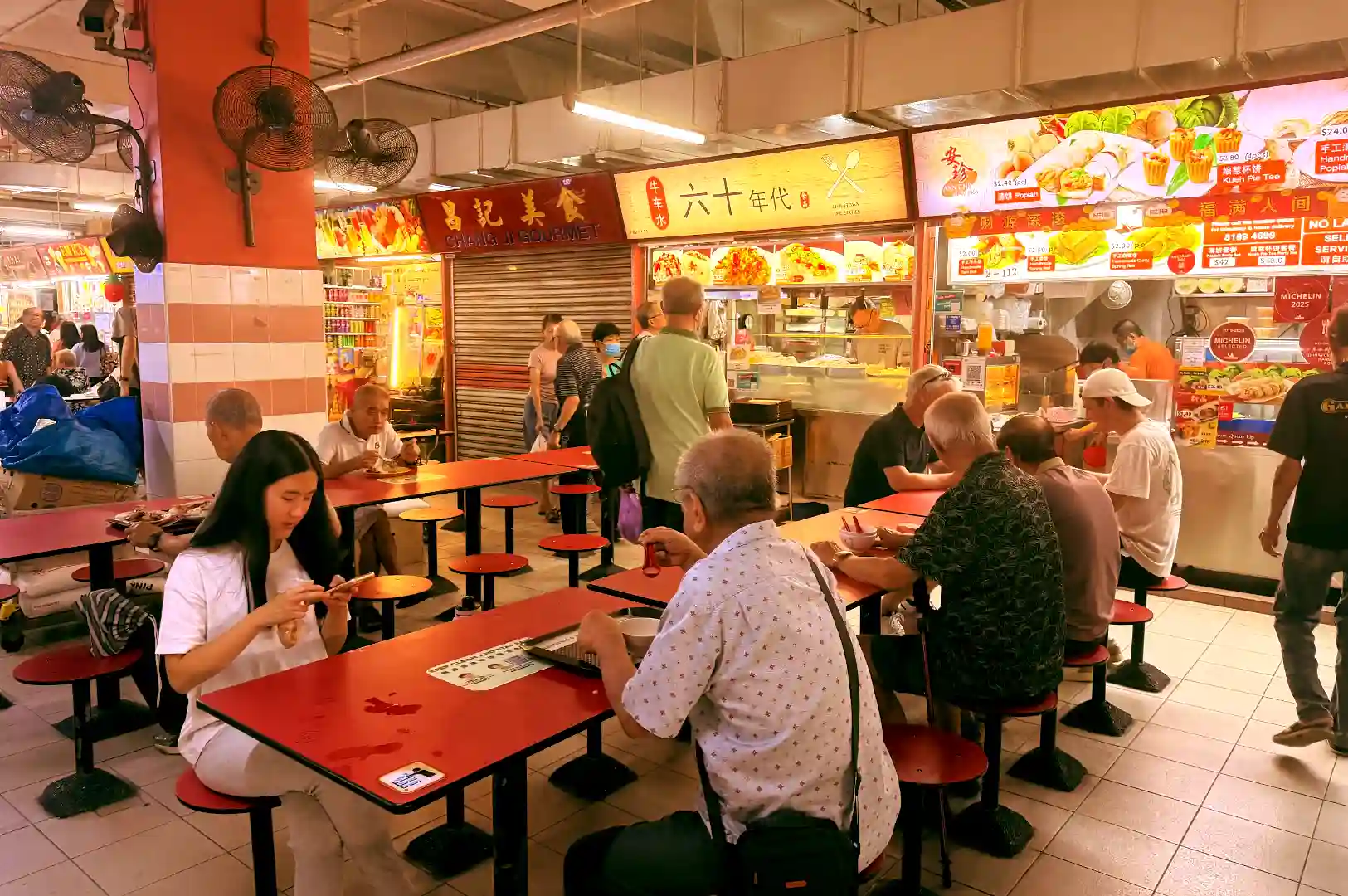 Unposed candid shot of diners seated at red tables inside a bustling Singapore hawker centre during peak meal hours.