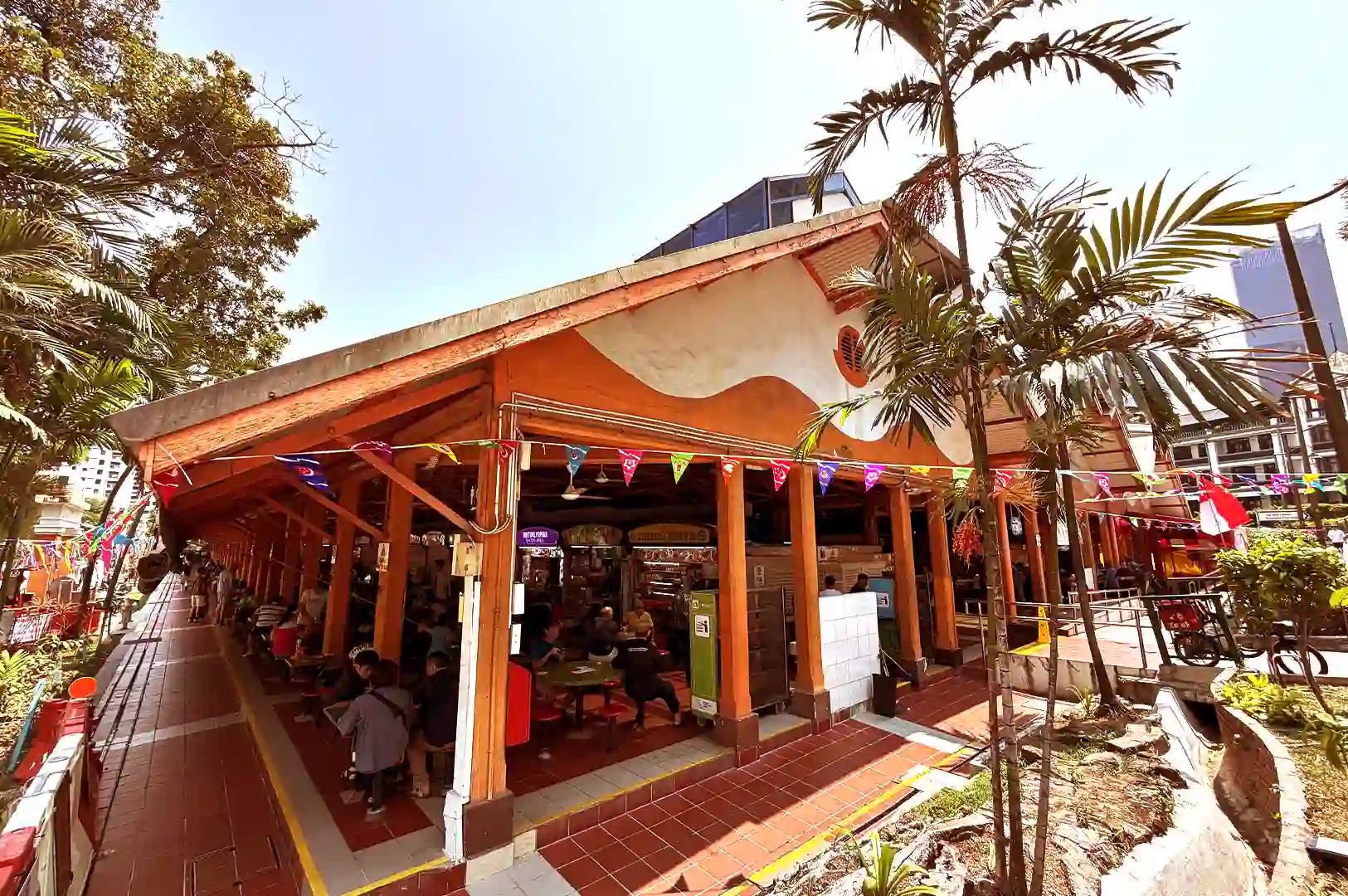 Exterior view of an open-air Singapore hawker centre under an orange wooden roof structure surrounded by palm trees and colorful bunting decorations.