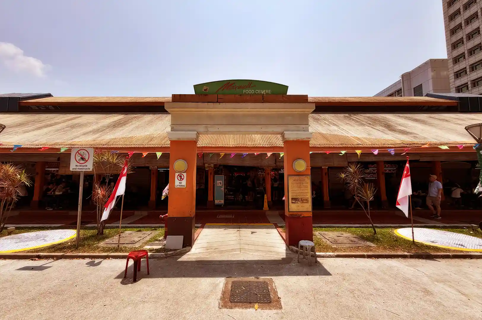 The heritage entrance archway of Maxwell Food Centre under a bright blue sky, framing its historic corrugated roof and market origins where generations have gathered.