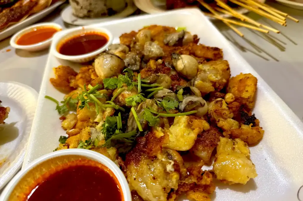 Close-up of crispy fried oyster omelette topped with juicy oysters and fresh coriander, served on a plate alongside spicy chili dip at Newton Food Centre.