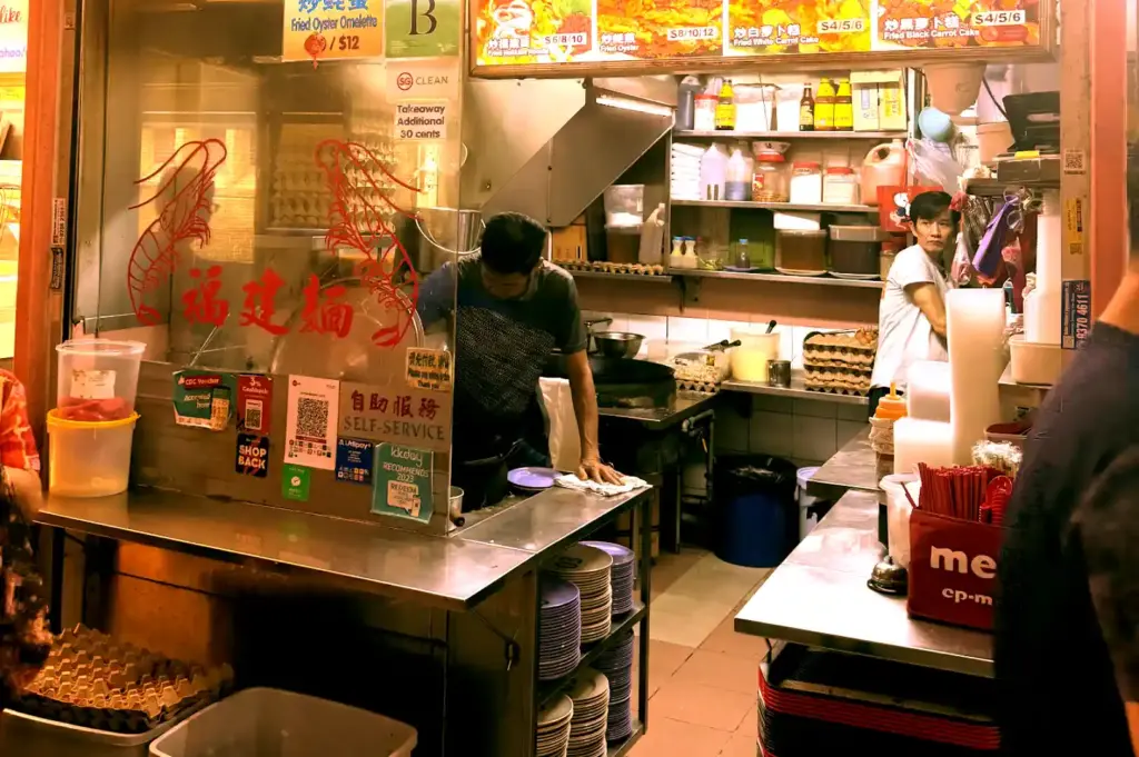 Behind-the-scenes view of a hawker stall kitchen filled with stacked plates, egg crates, and a cook cleaning the counter during service.