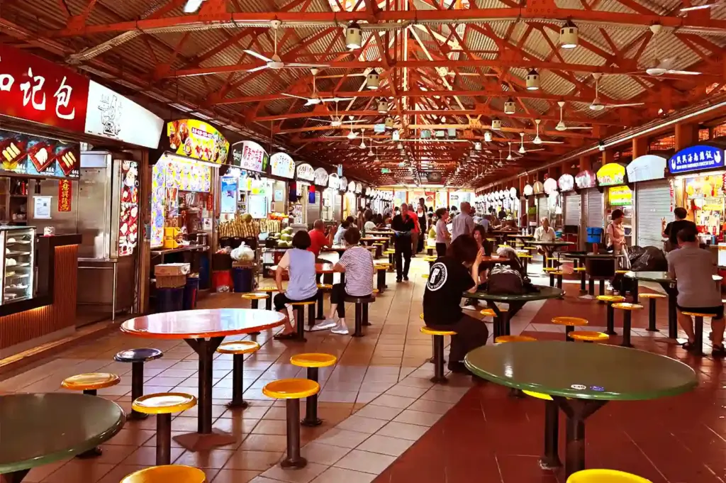 Bustling interior of a traditional hawker food centre with seated diners, vibrant food stall signboards, and high exposed ceiling beams.