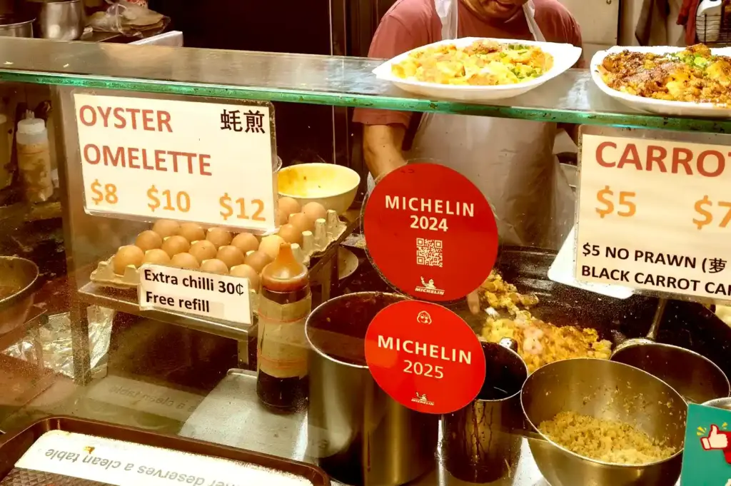 A hawker cook frying carrot cake on a large flat-top griddle behind a counter displaying Michelin Bib Gourmand stickers and pricing signs.