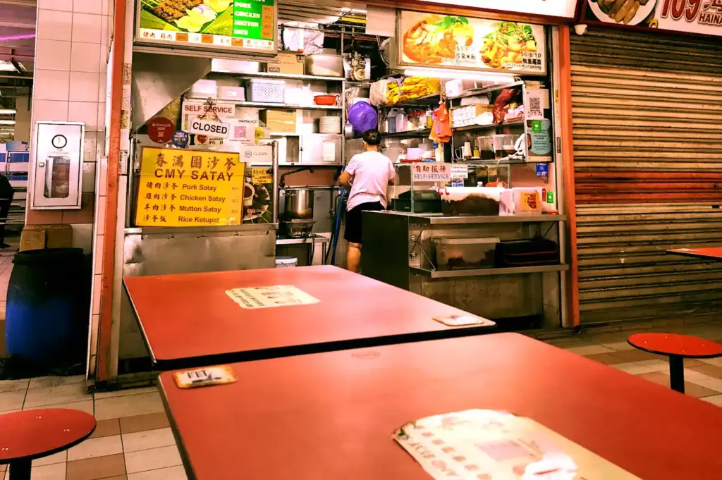 A quiet moment at a closed CMY Satay stall as a hawker worker cleans the kitchen area behind empty dining tables