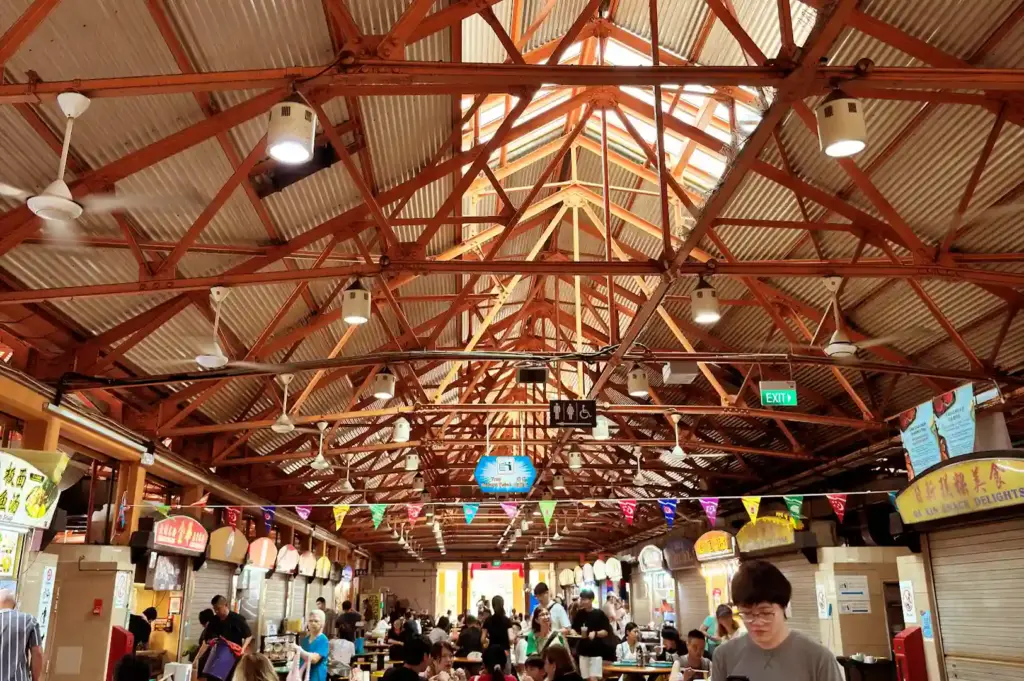 Looking up at the expansive, intricate steel truss architecture and hanging lights of Maxwell Food Centre’s cavernous ceiling, where the tables remember decades of shared meals.